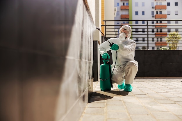 Mann in steriler Schutzkleidung hockt und besprüht die Wand mit Desinfektionsmittel, um die Ausbreitung des Coronavirus zu verhindern.