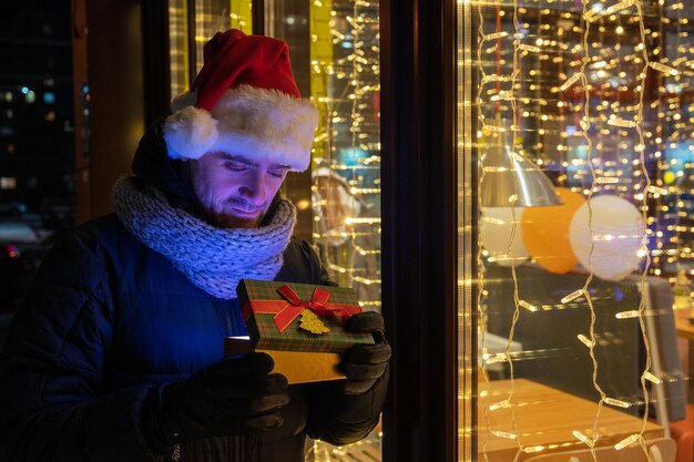 Mann in Weihnachtsmütze mit Geschenkbox in der Nähe des beleuchteten Café-Fensters