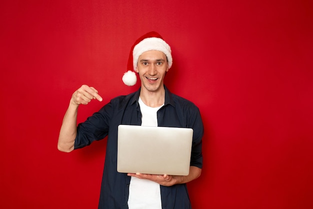 Mann in Weihnachtsmütze zeigt mit seinem Zeigefinger-Laptop in seinen Händen isoliert auf rotem Hintergrund
