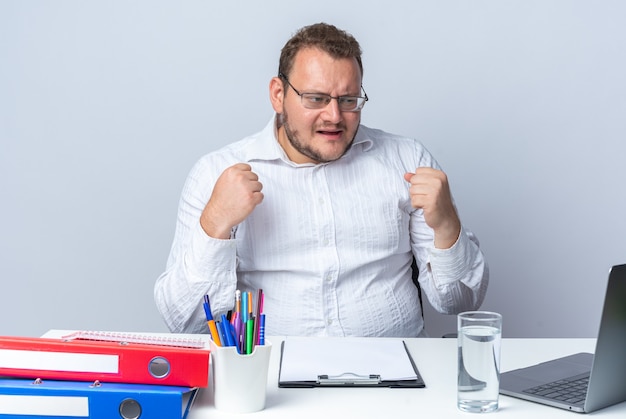 Mann in weißem Hemd mit Brille, der am Tisch mit Büroordnern und Zwischenablage sitzt und auf den Bildschirm des Laptops schaut, glücklich und aufgeregt, die Fäuste über der weißen Wand zusammenballt, die im Büro arbeitet