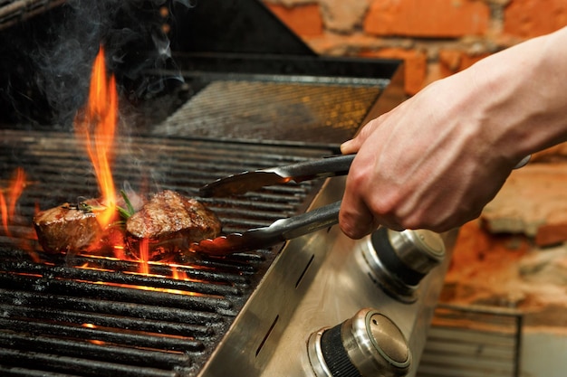 Mann kocht Grillfleischsteaks auf professionellem Grill im Freien. Männliche Hand mit Zange, die Beefsteaks auf offenem Feuer dreht