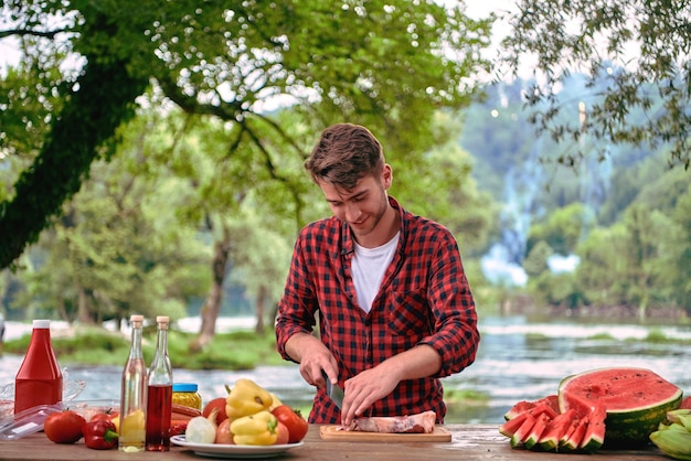 Mann kocht leckeres Essen auf dem Grill für eine französische Dinnerparty im Freien in der Nähe des Flusses an einem schönen Sommerabend in der Natur