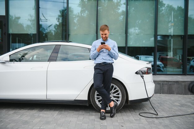 Mann lädt sein Elektroauto an Ladestation und nutzt Smartphone and