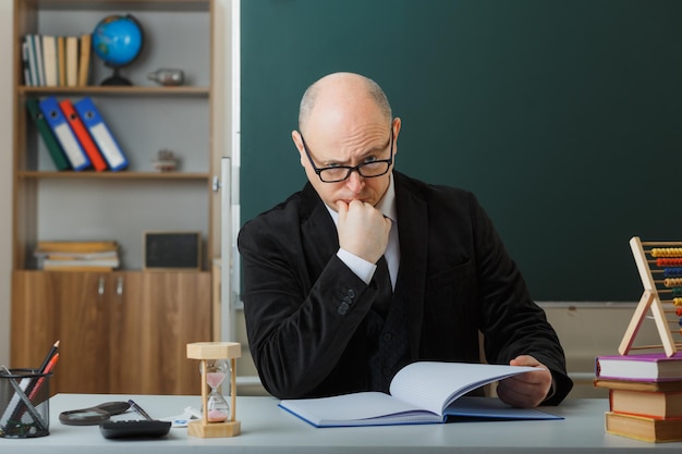 Mann, Lehrer, der eine Brille trägt, die das Klassenbuch überprüft und in die Kamera schaut, denkt stirnrunzelnd, wie er an der Schulbank vor der Tafel im Klassenzimmer sitzt