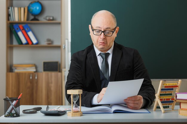 mann, lehrer, der eine brille trägt, sitzt an der schulbank vor der tafel im klassenzimmer und überprüft die hausaufgaben der interessierten schüler