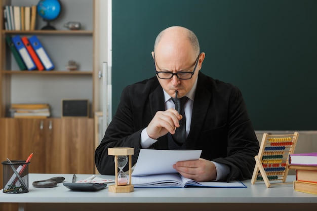 Mann Lehrer mit Brille sitzt an der Schulbank vor der Tafel im Klassenzimmer und überprüft die Hausaufgaben der Schüler konzentriert mit ernster Miene