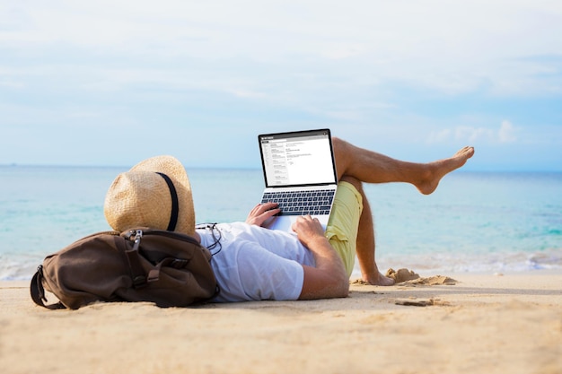 Mann liest E-Mails auf Laptop, während er sich am Strand entspannt