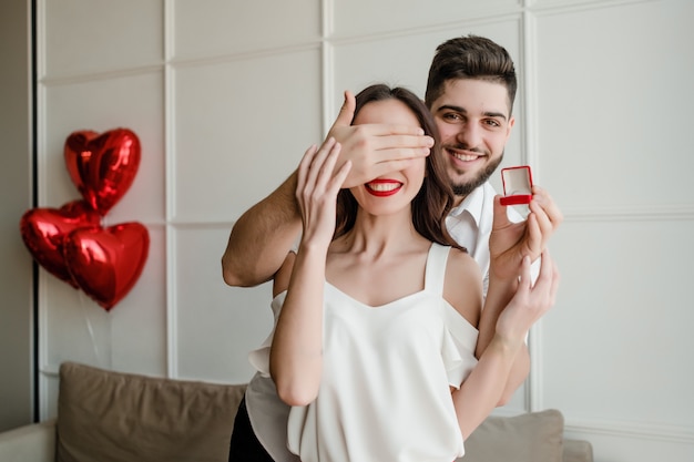 Mann macht der Frau mit Ring zu Hause mit Herz geformten Ballonen auf Couch in der Wohnung Überraschungsantrag