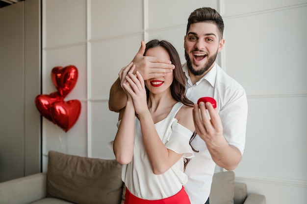 Mann macht der Frau mit Ring zu Hause mit Herz geformten Ballonen auf Couch in der Wohnung Überraschungsantrag