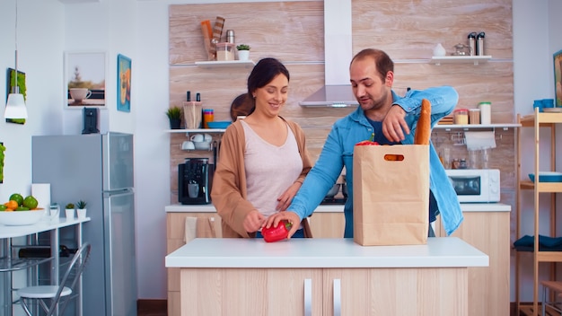 Mann macht Tricks mit Orangen in der Küche, während Frau Gemüse aus Papiertüte in den Kühlschrank legt. Fröhlicher, glücklicher, gesunder Lebensstil der Familie, frisches Gemüse und Lebensmittel. Supermarkt produ