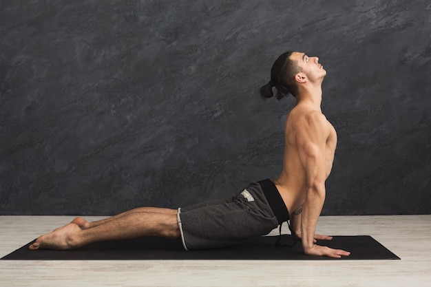 Mann macht Yoga. Sportler, der eine Cobra-Haltung macht, sich auf der Matte auf grauem Hintergrund zurückstreckt, Platz kopieren