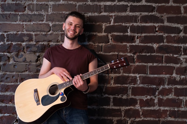 Mann mit akustischer Gitarre gegen Backsteinmauer, die Musik spielt, Lieder singen, das Leben genießen Hübscher kaukasischer männlicher Gitarrist, der übt, Musikinstrument zu Hause im Loft-Interieur zu spielen Kreatives Lifestyle-Hobby