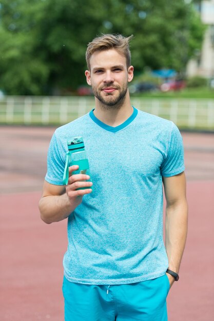 Mann mit athletischem Aussehen hält Flasche mit Wasser. Sportler trinken Wasser nach dem Training im Stadion an einem sonnigen Tag. Bemannen Sie Sportler in sportlicher Kleidung, die im Freien trainiert. Sport und gesundes Lifestyle-Konzept.