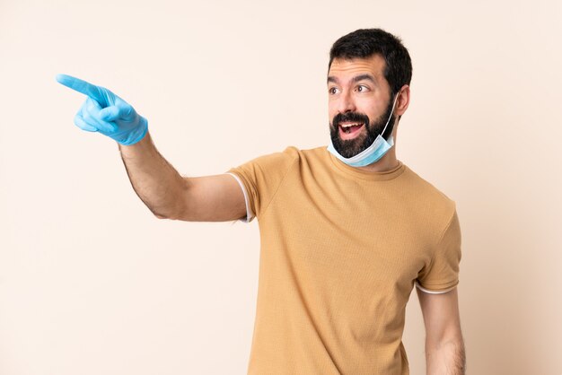 Mann mit Bart, der vor dem Coronavirus mit einer Maske und Handschuhen über isolierter Wand schützt, die weg zeigt