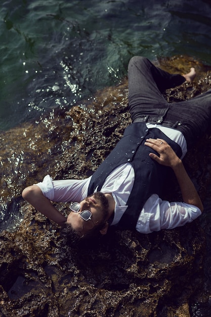 Mann mit Bart in dunkler Kleidung und weißem Hemd liegt an der steinernen Küste auf der Krim tarkhankut