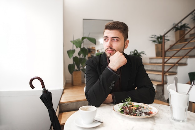 Mann mit Bart und Anzug sitzt in einem Restaurant mit Salat und einer Tasse Kaffee auf dem Tisch und schaut aus dem Fenster