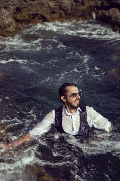 Mann mit Bart und Sonnenbrille in Kleidung, Weste und weißem Hemd schwimmt im Sommer zwischen den Felsen im Meer