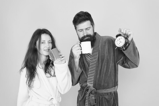 Mann mit Bart und verschlafene Frau genießen Morgenkaffee oder Tee Zeit zum Aufwachen und schönen Tag haben Guy Ehemann hält Kaffee und Uhr Jeder Morgen beginnt mit Kaffee Paar in Bademänteln mit Tassen