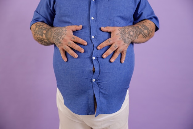 Mann mit Übergewicht und Tätowierungen hält die Hände auf dem großen Bauch auf violettem Hintergrund