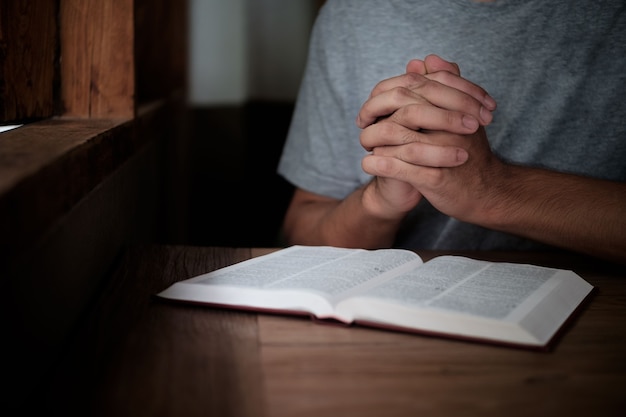 Mann mit Bibel, die betet, Hände auf ihrer Bibel auf Holztisch gefaltet.