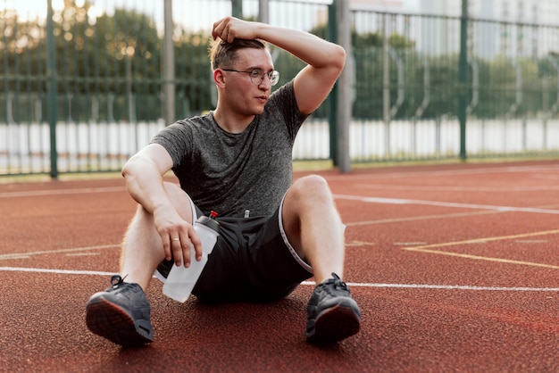 Mann mit Brille, der Sportkleidung trägt, sitzt am Basketballplatz und hält eine Flasche mit Wasser in den Händen