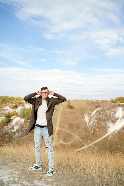 Mann mit Brille in Jeans T-Shirt und Umhang steht in hügeligem Gelände Erwachsener Mann in Kapuze passt seine Brille an und genießt bei windigem Wetter Zeitlupe einen schönen Blick auf die Landschaft