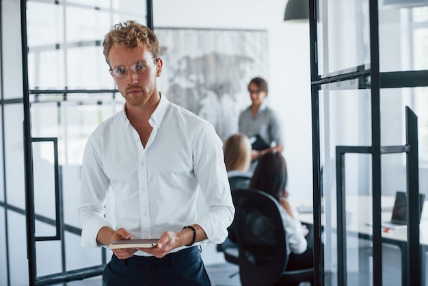 Mann mit Brille mit Notizblock in den Händen steht im Büro gegenüber Mitarbeitern.