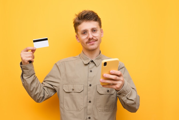 Mann mit Brille und Schnurrbart hält Kreditkarte und Smartphone, schaut in die Kamera und lächelt gegen Gelb