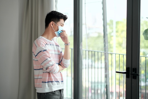 Mann mit chirurgischer maske, der aus seinem hausfenster schaut. Konzept für Coronavirus, Covid-19 und Quarantäne.