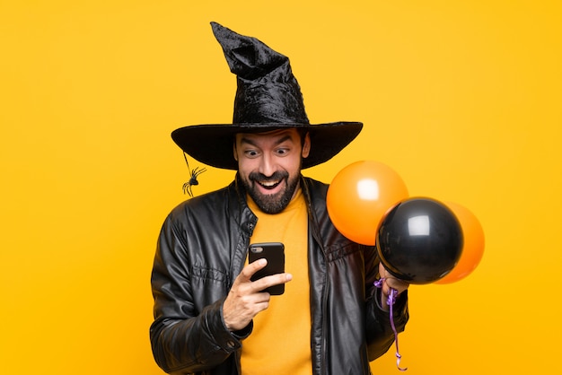 Mann mit dem Hexenhut, der die schwarzen und orange Luftballone für Halloween-Partei überrascht hält und eine Mitteilung sendet