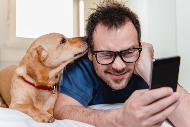 Mann mit dem Hund, der intelligentes Telefon auf dem Bett verwendet