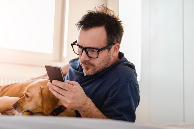 Mann mit dem Hund, der intelligentes Telefon auf dem Bett verwendet