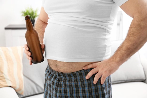Mann mit dickem Bauch, der zu Hause eine Flasche Bier hält