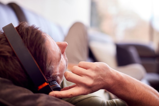 Mann mit drahtlosen Kopfhörern, die Musik hören, die zu Hause auf dem Sofa liegt und einen Schlafanzug trägt