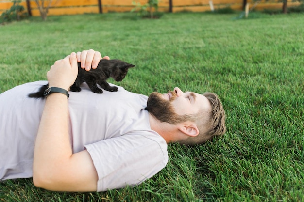 Mann mit einem kleinen Kätzchen, das auf dem Gras liegt und spielt, Freundschaft, Liebe zu Tieren und Konzept des Haustierbesitzers