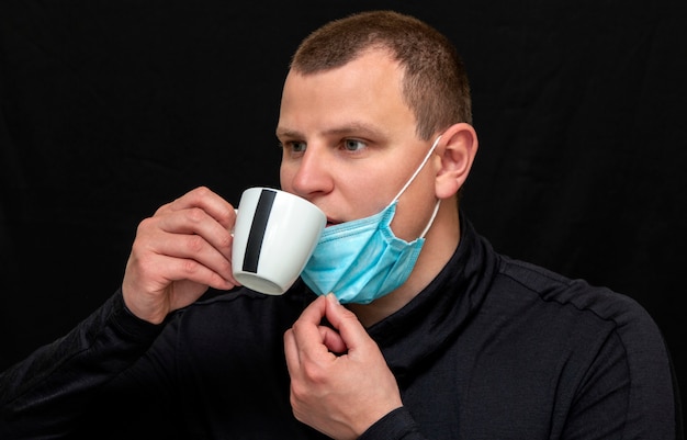 Mann mit einer medizinischen Maske trinkt Kaffee an einer dunklen Wand