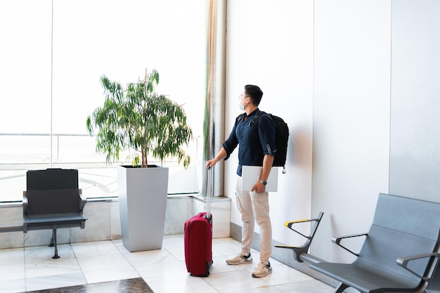 Mann mit Gesichtsmaske, der darauf wartet, an Bord zu gehen Kerl mit Koffertasche und Computer-Laptop, der am Flughafen steht