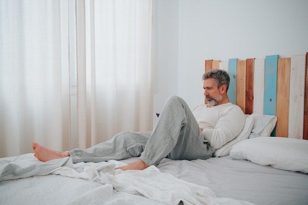Mann mit grauen Haaren in den 50ern, der auf seinem Bett liegt und beim Aufstehen auf das Telefon schaut