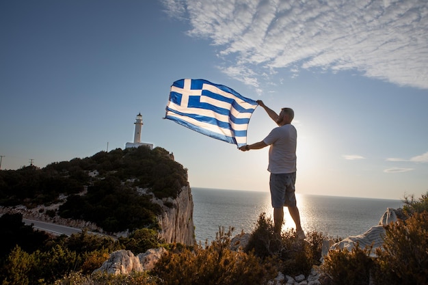 Mann mit griechischer Flagge bei Sonnenuntergang über dem Meer Leuchtturm der Insel Lefkada im Hintergrund