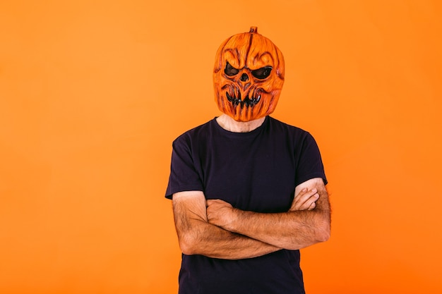 Foto mann mit gruseliger kürbislatexmaske mit blauem t-shirt mit verschränkten armen, auf orangem hintergrund. halloween und tage des toten konzepts.
