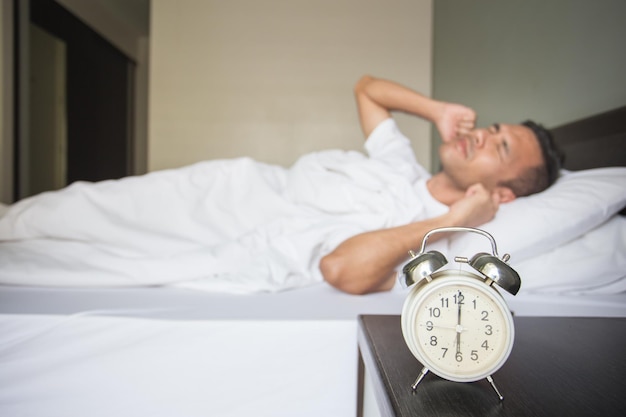 Foto mann mit händen, die die ohren über dem bett bedecken, mit wecker am nachttisch im schlafzimmer