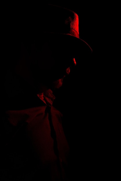 Mann mit Hut auf dunklem Hintergrund, Sonnenbrille und rotem Licht im Gesicht. Studioporträt