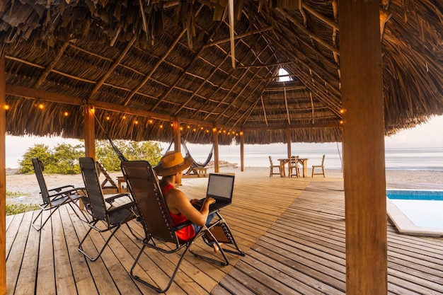 Mann mit Hut sitzt auf einem Stuhl und arbeitet an einem Laptop vor dem Strand