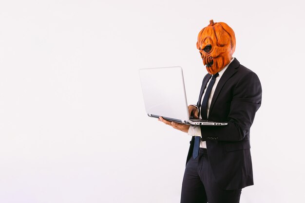 Mann mit Jackenanzug, blauer Krawatte und Jack-o-Laternen-Kürbismaske, der mit seinem Laptop arbeitet. Halloween- und Karnevalsfeierkonzept.