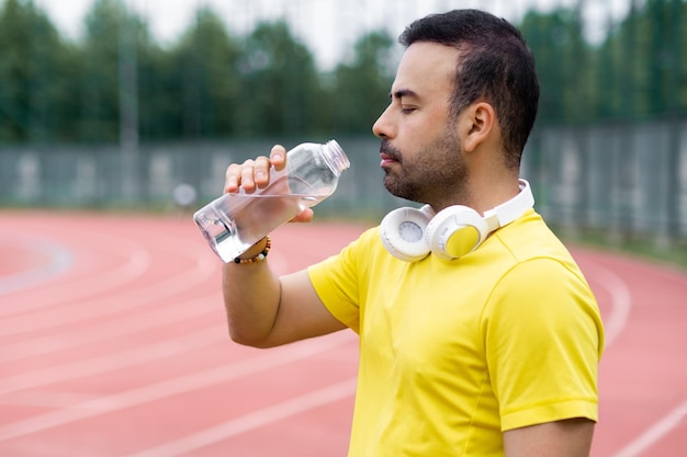 Mann mit kabellosen Kopfhörern blickt in offene Wasserflasche und ruht sich nach dem Training auf der Laufstrecke aus