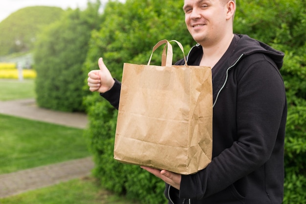 Mann mit Öko-Papiertüte für Speisen zum Mitnehmen mit Daumen nach oben auf naturgrünem Hintergrund Lieferung bei jedem Wetter rund um die Uhr an den Kunden