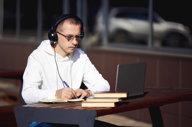 Mann mit Kopfhörern arbeitet an einem Laptop und schreibt in ein Notizbuch, das auf der Straße an einem Tisch sitzt