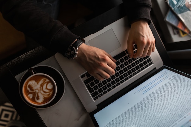 Mann mit Laptop und Kaffee arbeitet und tippt drinnen auf der Tastatur. Männliche Hände mit Luxusuhr, die in einem Café arbeitet