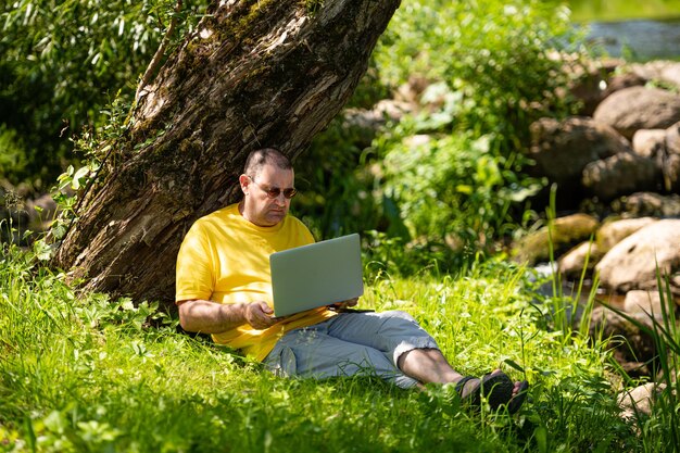 Mann mit Laptop und Smartphone, der draußen auf der Wiese am Fluss-Bürokonzept arbeitet