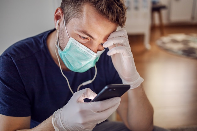 Mann mit Maske und Handschuhen, die Musik hören und am Smartphone tippen. Bleib zuhause. Weltpandemie, Coronavirus.